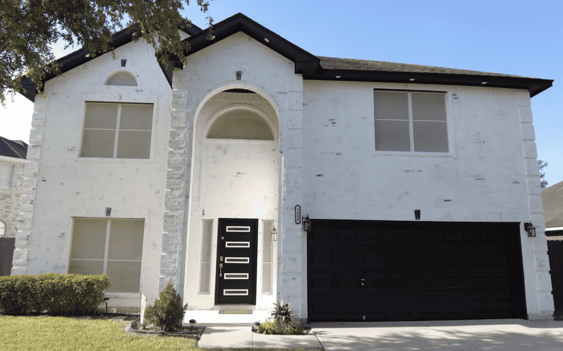 Two-story white brick house with arched entryway, modern black door, and matching garage. Trimmed hedges and a neatly cut lawn in the foreground.